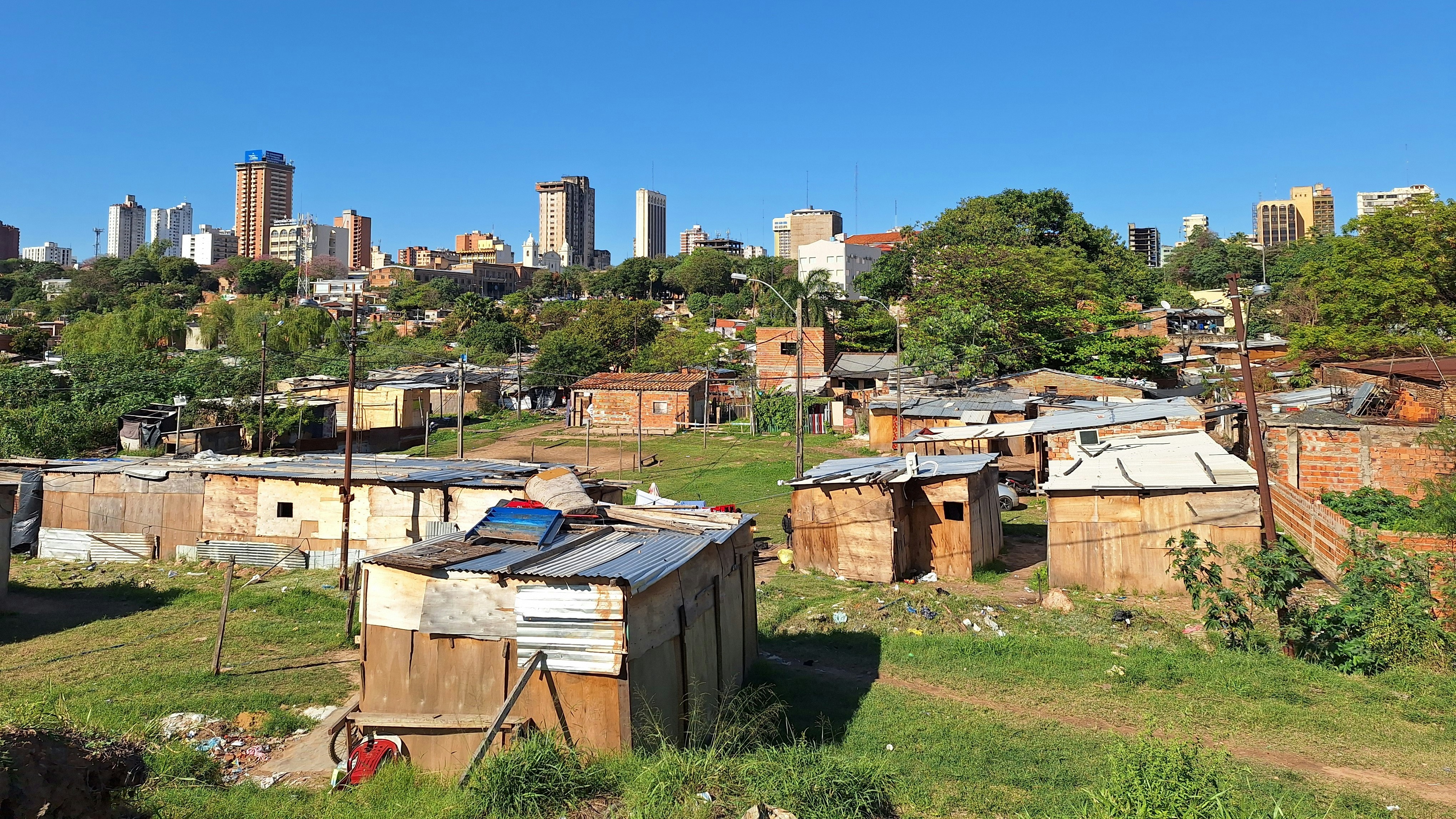 Informal settlements in the foreground with metal roofs, and a modern skyline in the background; contrast between poverty and urban development.