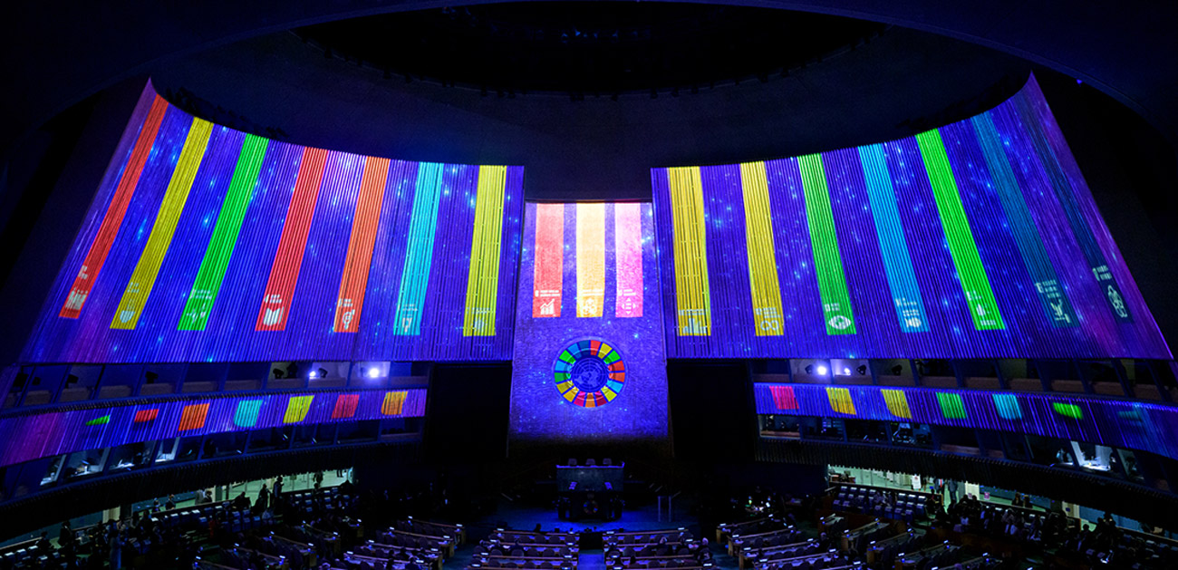 Sala de conferencias con paneles en el fondo y costados, donde se ven reflejadas franjas con el color de cada ods. Al centro está el círculo con los colores de los 17 Objetivos del Desarrollo Sostenible